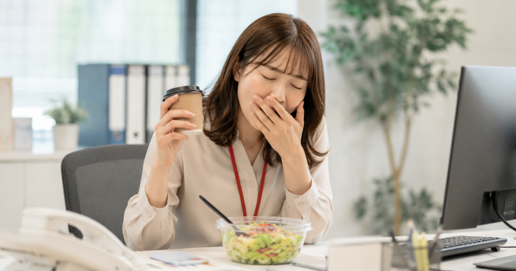昼食後にあくびをするオフィス女性。食後眠い・だるいという悩みに共感できるリアルなシーン。